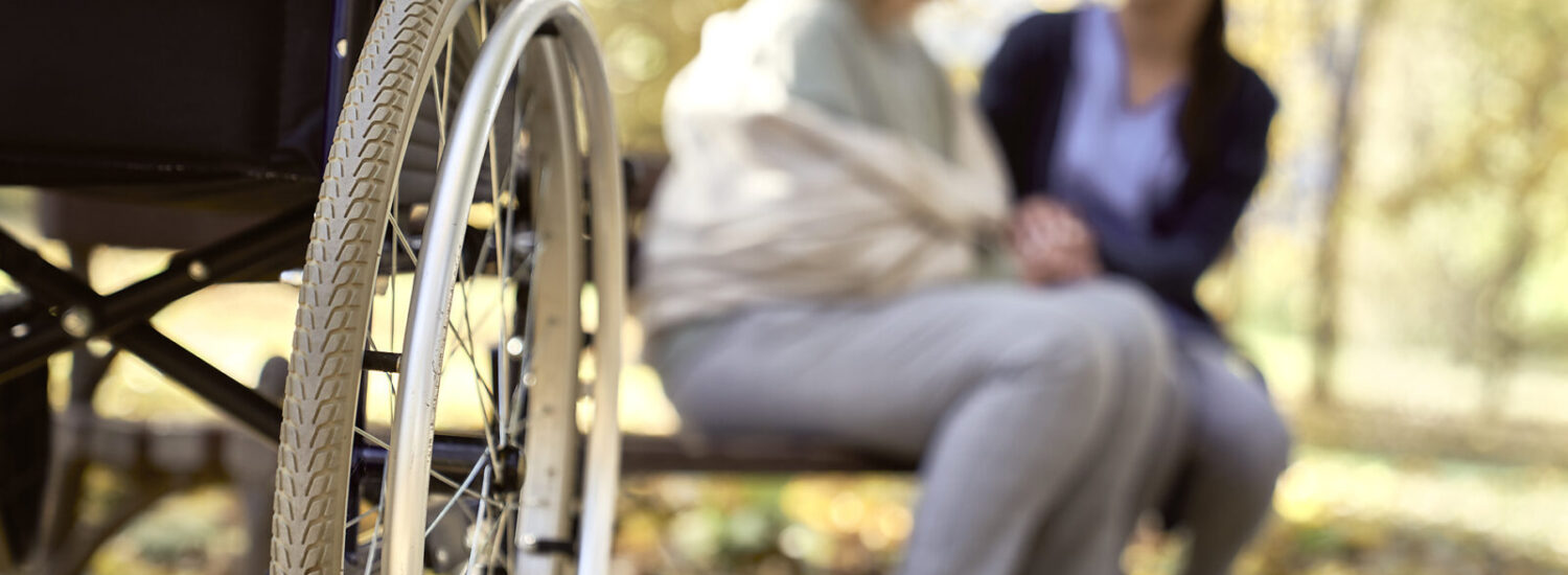 Rollstuhl mit zwei Menschen auf Bank im Hintergrund © Westend61 / gpointstudio