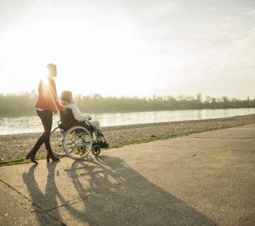 Frau schiebt andere Frau in Rollstuhl an einem Fluss entlang © Westend61 / Uwe Umstätter