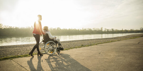 Frau schiebt andere Frau in Rollstuhl an einem Fluss entlang © Westend61 / Uwe Umstätter