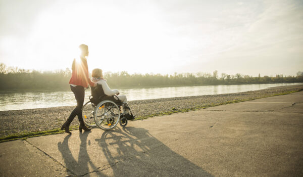 Frau schiebt andere Frau in Rollstuhl an einem Fluss entlang © Westend61 / Uwe Umstätter Frau schiebt andere Frau in Rollstuhl an einem Fluss entlang © Westend61 / Uwe Umstätter