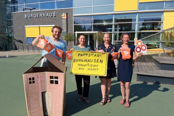 2025_07_14_Stadt_Burghausen_PM_Pappstadt Ein Gebäude gibt es schon, sie hoffen auf viele weitere: Die Initiatoren der Bastelaktion „Pappstad“ (v.l.) Marcel Anslinger (Leiter Freizeitheim), Mariella Röben (Kinderschutzbund), Simone Mayer (Werbering) und Monika Lengyel (Freizeitheim).