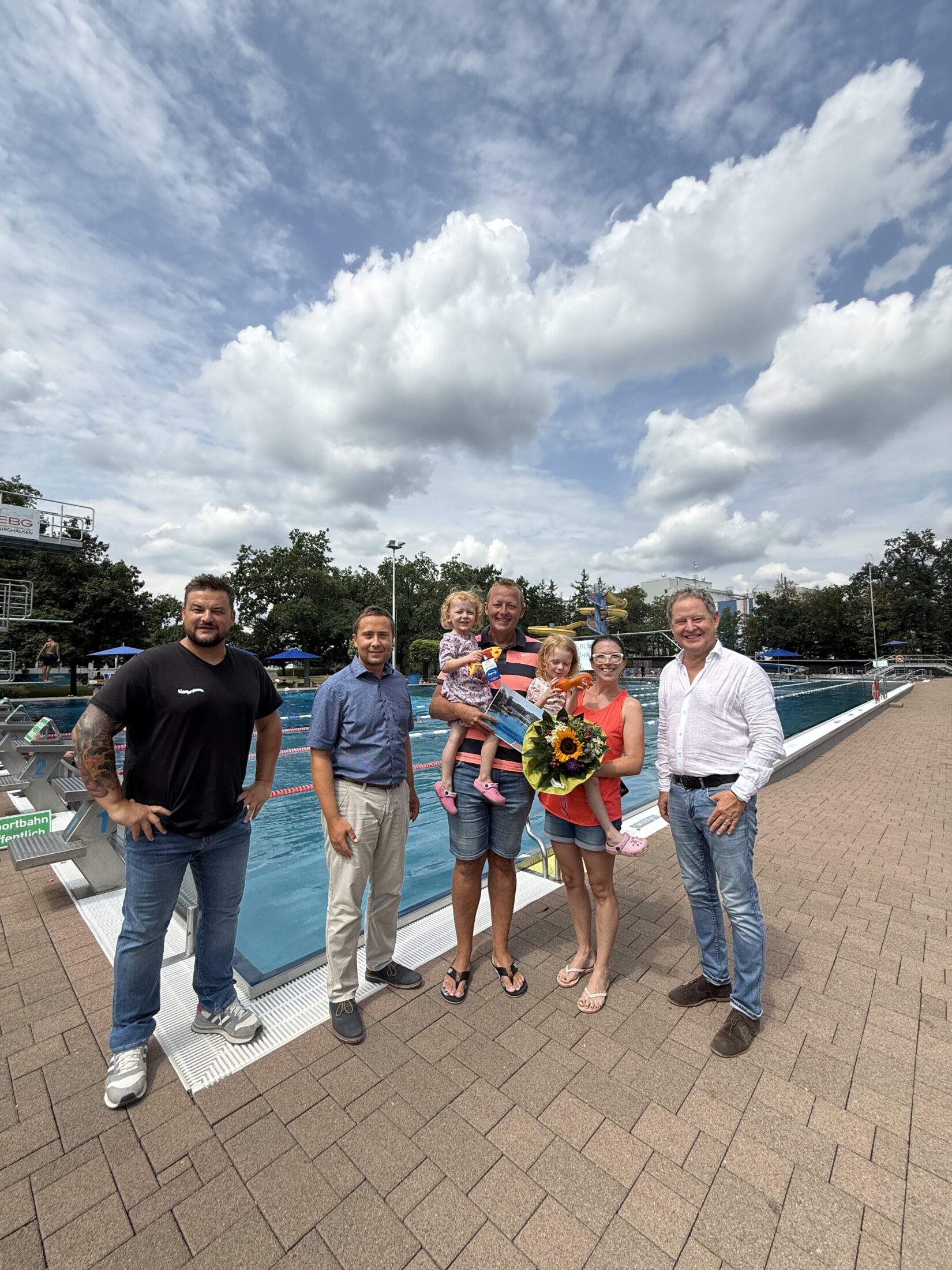 Erster Bürgermeister Florian Schneider begrüßte gemeinsam mit Bäderleiter Stefan Sajdak und Florian Moser die Familie Kornberger aus Ampfing als 100 000  Badegäste der Freibadsaison 2026 im Burghauser Freibad  Zur Überraschung gab es Blumen für Susanne Kornberger, Taucherbrillen für Teresa und Valentina sowie eine Saisonkarte 2027 für die ganze Familie  Fotocredit: Stadt Burghausen / köx
